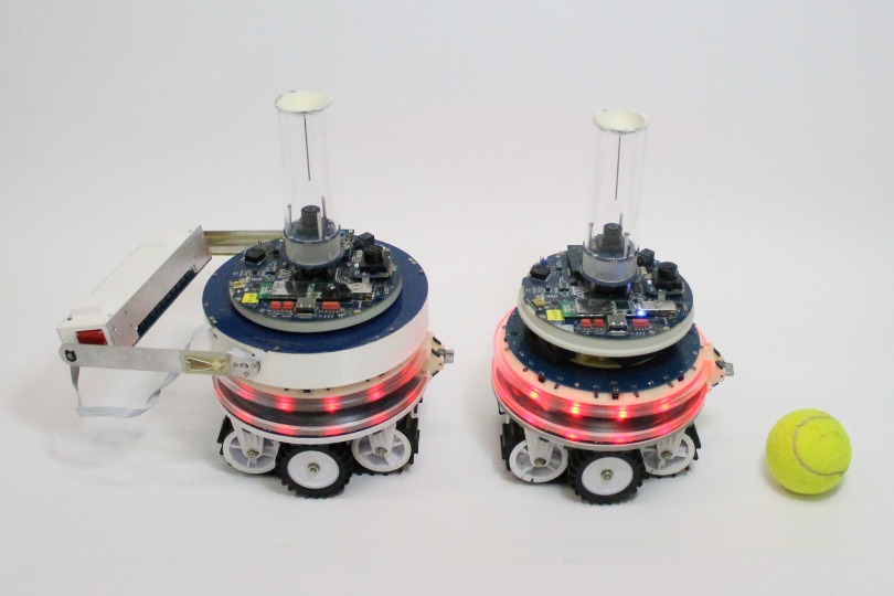 Two autonomous MNS robots in different configurations shown alongside a tennis ball for scale. Left: hardware configuration with object lifting capabilities. Right: standard configuration.